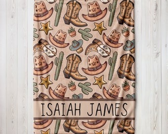 May include: A tan blanket featuring a repeating pattern of cowboy hats, boots, cacti, and stars. The blanket has a brown band with the name "ISAIAH JAMES" printed in black.