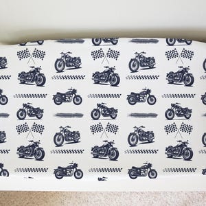 May include: White fabric with a repeating pattern of blue motorcycles and checkered flags. The motorcycles are all facing the same direction and have a classic design.