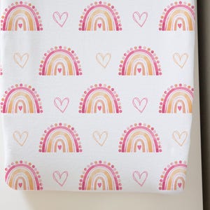 May include: White fabric with a repeating pattern of watercolor rainbows and hearts in shades of pink, orange, and peach. The rainbows have a dotted top border. The design is suitable for baby items.