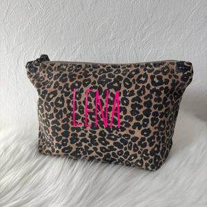 Könnte beinhalten: Eine Kosmetiktasche mit Leopardenmuster und dem Namen "LENA" in leuchtendem Pink gestickt. Die Tasche hat einen Reißverschluss und ist vor einem weißen, flauschigen Hintergrund platziert.