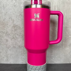 May include: A pink stainless steel travel mug with a black and white patterned base. The mug has a handle and a lid with a straw. The mug has the Stanley logo on the front.