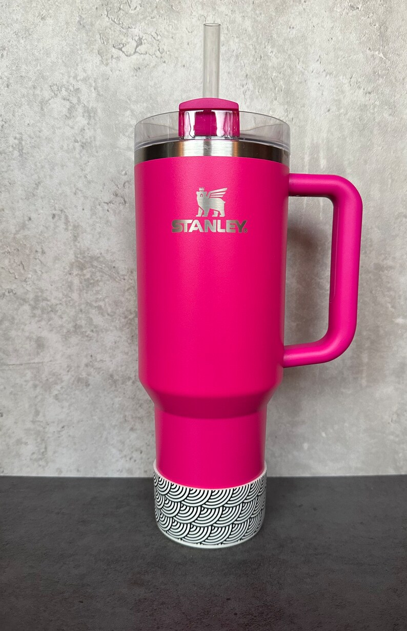 May include: A pink stainless steel travel mug with a black and white patterned base. The mug has a handle and a lid with a straw. The mug has the Stanley logo on the front.