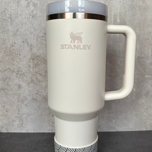May include: White Stanley travel mug with a black and white patterned band around the bottom. The mug has a handle and a lid with a straw hole. The Stanley logo is printed on the side of the mug.
