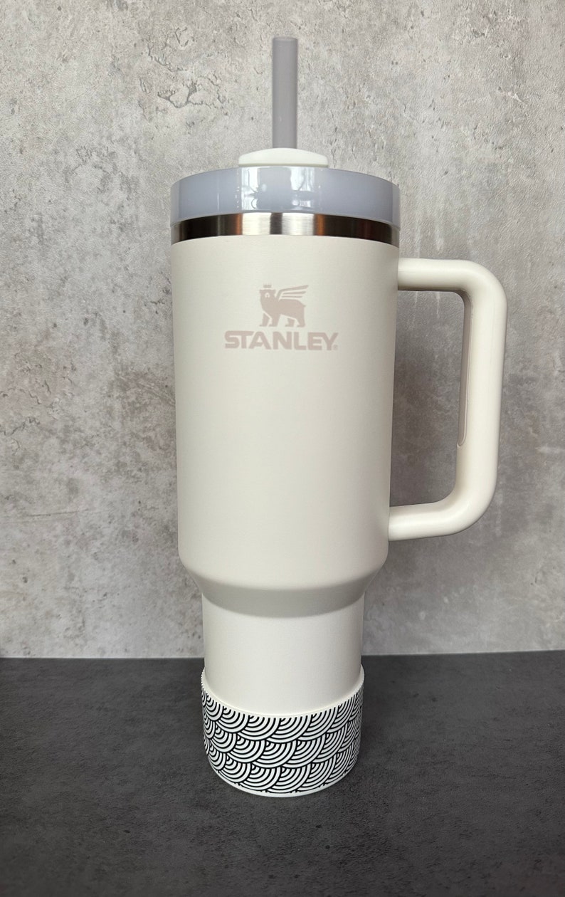 May include: White Stanley travel mug with a black and white patterned band around the bottom. The mug has a handle and a lid with a straw hole. The Stanley logo is printed on the side of the mug.