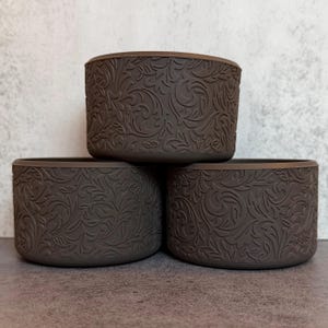 May include: Three dark brown cylindrical containers stacked in a pyramid formation. Each container features a continuous, embossed, swirling floral and leaf pattern covering the entire exterior surface, creating a textured, matte appearance.