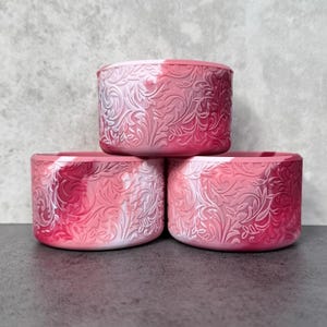May include: Three stacked, cylindrical planters in shades of pink and white. Each planter features an embossed floral design. The planters are arranged on a dark surface against a gray background.
