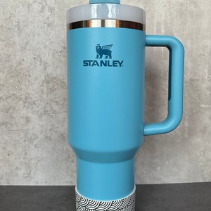 May include: A light blue stainless steel travel mug with a black and white patterned base. The mug has a handle and a lid with a straw hole. The mug has the Stanley logo and a bear with wings on it.