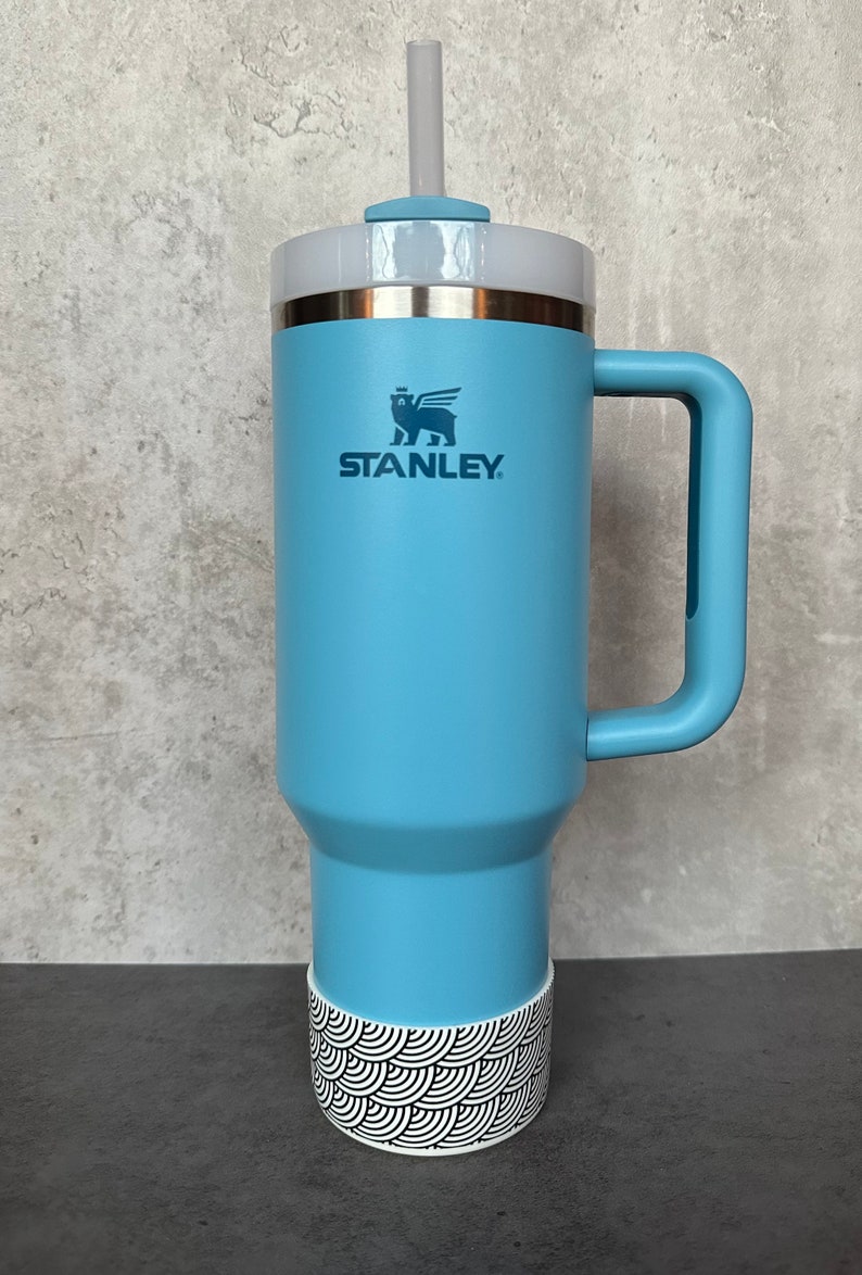 May include: A light blue stainless steel travel mug with a black and white patterned base. The mug has a handle and a lid with a straw hole. The mug has the Stanley logo and a bear with wings on it.