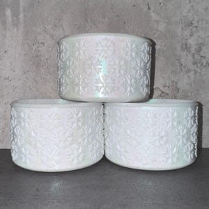 May include: Three white, cylindrical candle holders with a snowflake pattern. The holders are stacked, with the top one slightly angled. The snowflake design is embossed, and the holders have a pearlescent finish.