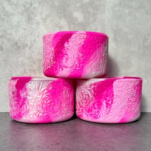 May include: Three stacked, cylindrical containers with a vibrant pink and white marbled design. Each container features an embossed floral pattern. The containers are made of a flexible material and are suitable for various uses.