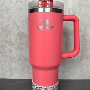 May include: A pink stainless steel travel mug with a black and white patterned base. The mug has a handle and a lid with a straw. The mug has the Stanley logo and the text "Since 1913".