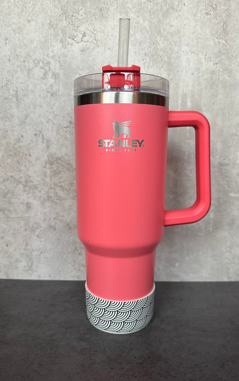 May include: A pink stainless steel travel mug with a black and white patterned base. The mug has a handle and a lid with a straw. The mug has the Stanley logo and the text "Since 1913".