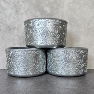 May include: Three silver-colored, cylindrical containers stacked. Each container features an embossed floral and leaf pattern. The containers are made of a metallic material and are displayed against a neutral background.