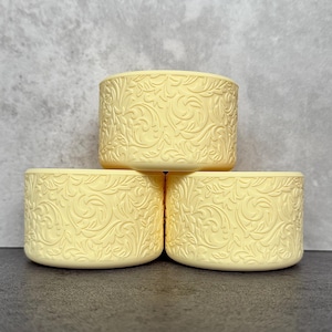 May include: Three stacked, pale yellow, cylindrical containers. Each container features an embossed floral and swirl pattern. The containers are arranged on a dark surface against a light gray background.