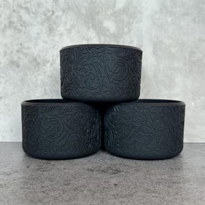May include: Three black, cylindrical water bottle sleeves with a detailed floral pattern. The sleeves are stacked, showcasing the embossed design. The background is a neutral gray, highlighting the dark color and texture of the sleeves.