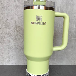 May include: A green stainless steel travel mug with a black and white patterned base. The mug has a handle and a lid with a straw. The mug has the Stanley logo on the front.