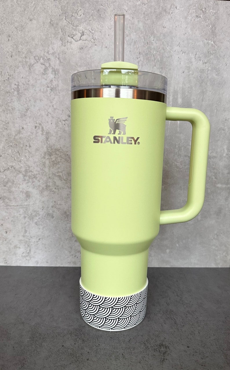 May include: A green stainless steel travel mug with a black and white patterned base. The mug has a handle and a lid with a straw. The mug has the Stanley logo on the front.