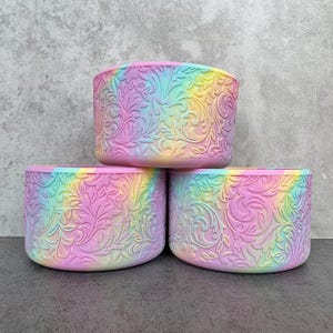 May include: Three cylindrical containers stacked, each with a pastel rainbow tie-dye effect. The containers feature an embossed floral pattern. The colors blend from pink to yellow to blue and green. The containers are made of a soft material.