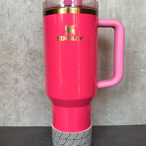 May include: A pink stainless steel travel mug with a black and white patterned base. The mug has a pink handle and a gold lid with a straw.