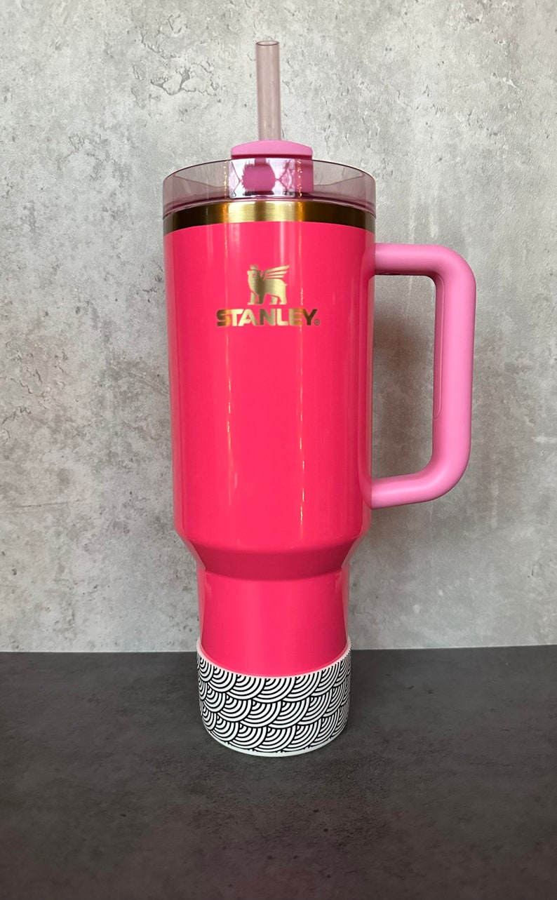 May include: A pink stainless steel travel mug with a black and white patterned base. The mug has a pink handle and a gold lid with a straw.