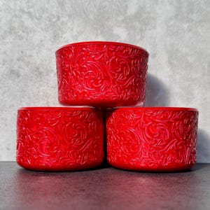 May include: Three stacked, cylindrical, red containers. Each container features an embossed, swirling floral pattern. The containers are arranged in a triangular formation, with the top container slightly offset.