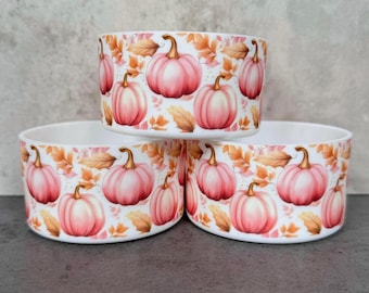May include: Three white ceramic bowls stacked, each adorned with a repeating pattern of pink pumpkins and autumn leaves. The pumpkins have brown stems, and the leaves are in shades of orange and brown. The bowls are cylindrical.