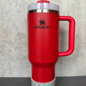 May include: A red Stanley travel mug with a black and white patterned base and a black logo that says "STANLEY". The mug has a handle and a lid with a straw.