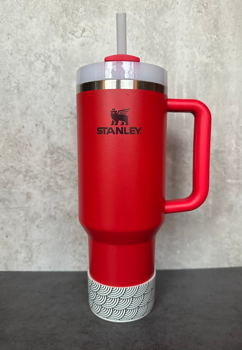 May include: A red Stanley travel mug with a black and white patterned base and a black logo that says "STANLEY". The mug has a handle and a lid with a straw.