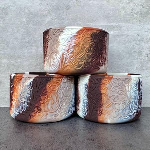 May include: Three decorative, cylindrical containers stacked. Each container features a repeating floral pattern in shades of brown, white, and orange. The containers are made of a material that appears to be silicone or a similar flexible substance.