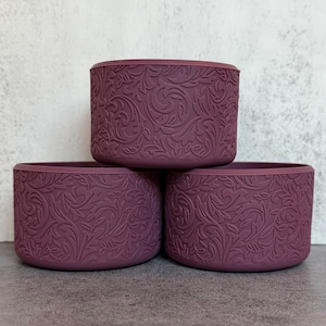 May include: Three cylindrical silicone boots in a deep plum color, each featuring a raised, intricate floral scrollwork pattern across the exterior surface. The items are arranged with one centered on top of two others, showcasing the texture.