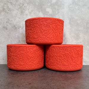 May include: Three vibrant orange cylindrical containers with an embossed floral pattern. The containers are stacked, showcasing the detailed design. The background is a neutral gray, highlighting the bright color and texture of the containers.