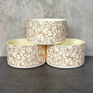 May include: Three white ceramic containers with a floral pattern in gold. The containers are stacked on top of each other.