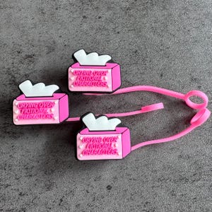 May include: Three pink shoe charms shaped like tissue boxes, each with white tissues and the text "Crying Over Fictional Characters." The charms are connected by a pink strap.
