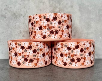 May include: Three pink ceramic bowls with a floral pattern. The bowls are stacked on top of each other. The pattern features white flowers, brown leaves, and orange accents.