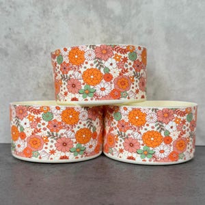 May include: Three white ceramic bowls with a floral pattern. The bowls have a white background with orange, pink, and green flowers.