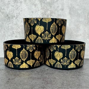 May include: Three black ceramic planters with gold leaf designs of various leaves. The planters are stacked on top of each other.