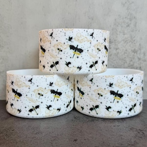 May include: Three white cylindrical bowls featuring a repeating pattern of black silhouettes of flying dragons, small gold-colored clouds, and tiny black stars scattered across the exterior surface of each container.