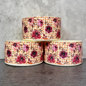 May include: Three cylindrical ceramic bowls stacked, each adorned with a floral pattern. The design features burgundy and pink roses, set against a tan and white gingham background. The bowls have a cream-colored rim.