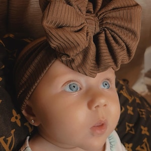 Peut inclure: Un bébé portant un bandeau en côtes brun avec un grand nœud sur le dessus. Le bébé porte un body blanc avec un imprimé vert et brun. Le bébé a les yeux bleus et regarde vers le haut.