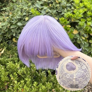 May include: A lavender-colored wig with a short, blunt cut and bangs. The wig features a lace cap with a floral design and small combs for secure attachment. The wig is displayed against a backdrop of green foliage.