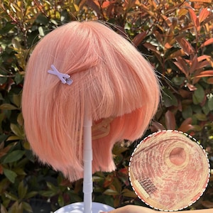 May include: A peach-colored wig with a short, layered bob cut. A small, light purple hair clip secures a section of the hair. The wig is displayed on a white stand, with a close-up showing the cap construction.