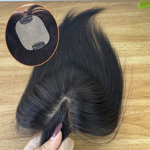 Op de afbeelding: Een donkerbruine haartop met een kanten basis en clips voor bevestiging. Het haarstuk wordt getoond op een houten oppervlak, met een close-up van de basis en clips. Het haar is gestyled om de scheiding te laten zien.
