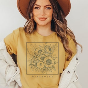 May include: Mustard yellow t-shirt featuring a square floral design with sunflowers and the word "Girasoles" in a serif font. The shirt is worn with a brown hat and a cream-colored jacket.