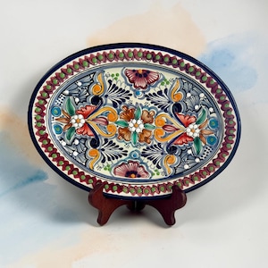 May include: An oval ceramic platter featuring a hand-painted floral pattern in shades of blue, yellow, green, and red. The border is decorated with a repeating pattern of small green dots and maroon petals, all set against a cream-colored background.