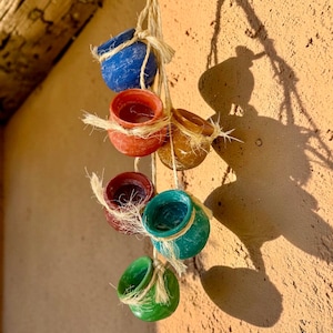 May include: A hanging mobile featuring six small, round, hollow clay pots in blue, orange, yellow, dark red, teal, and green. Each pot is individually tied with natural-colored fibrous twine, creating a vertical arrangement of colorful vessels.