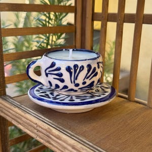 May include: A white ceramic teacup and saucer candle with a blue and white floral design. The candle is lit and the wick is visible.