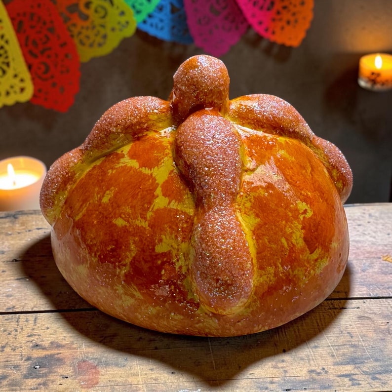 May include: A round, golden-brown pan de muerto bread featuring a central knob and four bone-shaped dough strips radiating from the top. The surface is glazed and sprinkled with fine sugar crystals, creating a textured, shiny appearance.