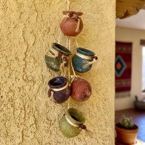 May include: A hanging cluster of small, colorful ceramic pots. The pots are in shades of red, green, blue, and purple, and are strung together with natural twine. Each pot is adorned with a twine bow and wooden beads.