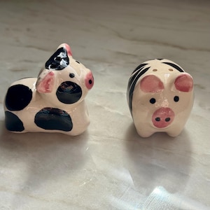 May include: Two ceramic pig-shaped salt and pepper shakers. One pig is white with black spots, the other is white with black stripes. Both have pink snouts and ears. The shakers are on a light-colored surface.
