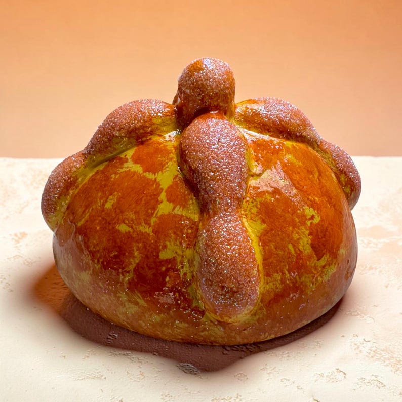 May include: A round, golden-brown pan de muerto bread featuring decorative dough strips arranged in a cross pattern on top. The surface of the baked bread is glazed and sprinkled with a fine layer of granulated sugar, creating a textured finish.
